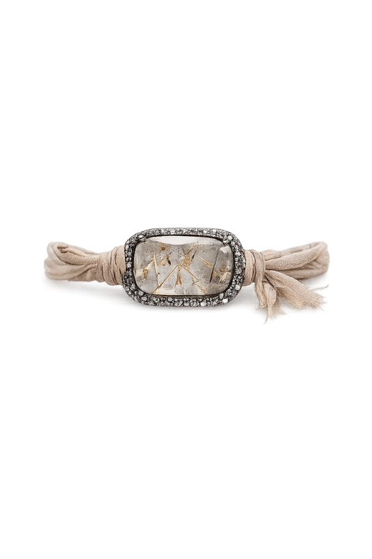 A beige fabric wrap bracelet with a central rectangular silver-toned bezel set with small clear stones and a textured, metallic gold-toned inlay, finished with knotted tassels on both sides.