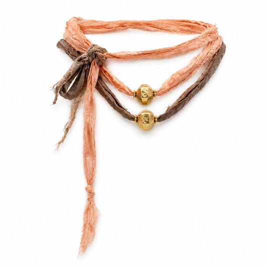 A bohemian-style multi-strand bracelet featuring peach-colored silk ribbon, dark brown woven cord, and two gold-colored metal beads, shown on a plain white background.