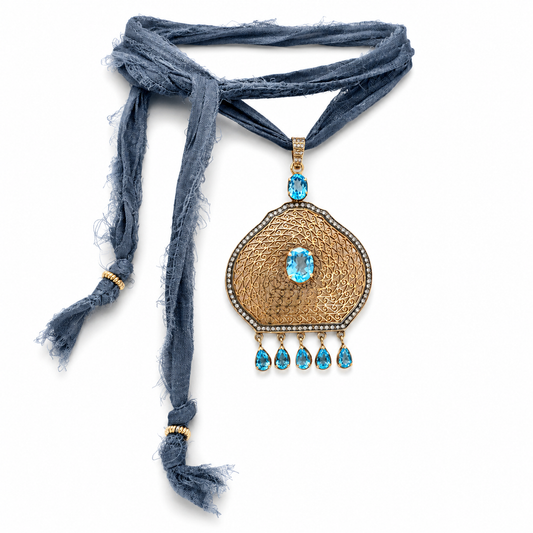 A bohemian-style necklace featuring a textured gold pendant with turquoise blue gemstones and a matching blue frayed scarf, displayed on a clean white background.