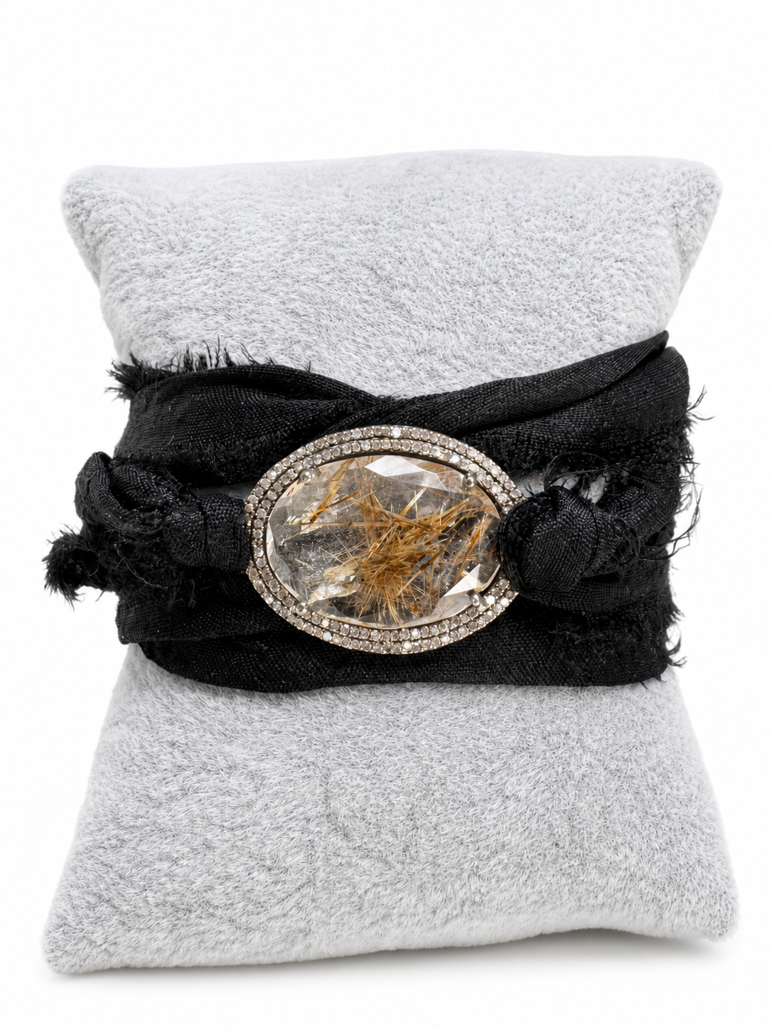 A black fabric wrap bracelet with a large oval clear crystal centerpiece containing golden inclusions, displayed on a light gray textured pillow against a white background.