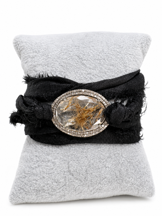 A black fabric wrap bracelet with a large oval clear crystal centerpiece containing golden inclusions, displayed on a light gray textured pillow against a white background.