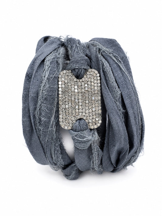 A distressed denim wrap bracelet in a faded blue-gray color, featuring a large rectangular silver-tone rhinestone-embellished clasp at the center, set against a plain white background.