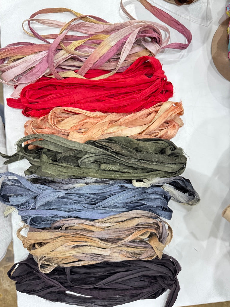 A vertical stack of multiple fabric ribbon or sash bundles in various colors, including pink, red, peach, olive green, blue, beige, and black, arranged on a white surface.