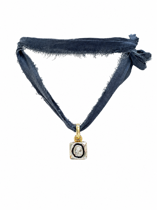 A dark blue fabric headband with frayed edges and a knotted side, featuring a gold pendant with a black oval center and a clear stone, shown on a plain white background.